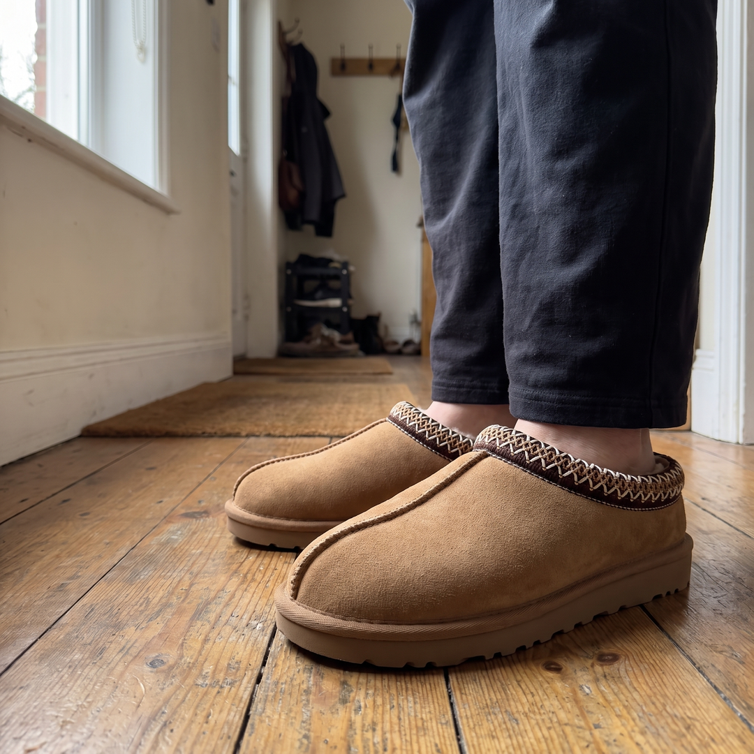 Pair of chestnut slip-on slippers worn indoors on a wooden floor with navy pants