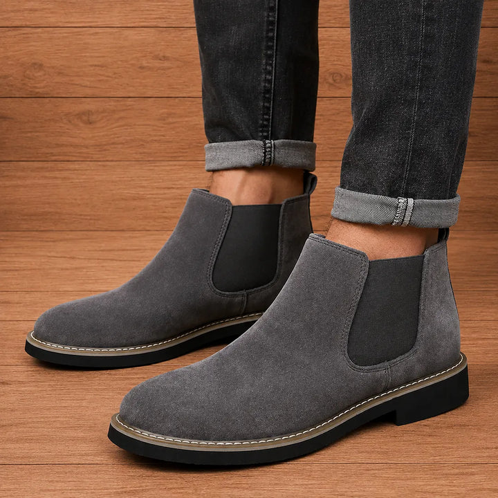 Dark gray chelsea boots worn with dark rolled jeans on a wood floor
