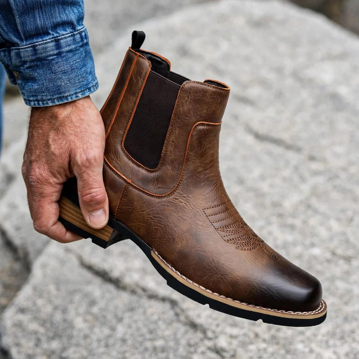 Hand holding a brown pull-on Chelsea boot against a rocky backdrop