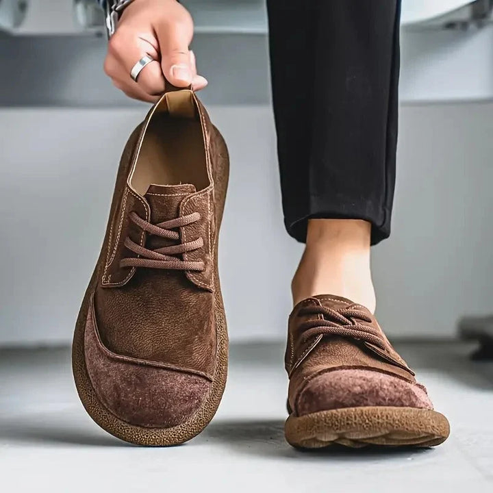 Brown wide toe lace up shoe held up beside a worn shoe with dark trousers