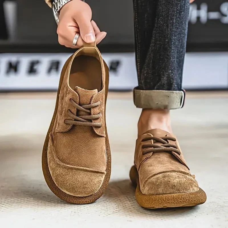 Tan wide toe lace up shoe held up beside a worn shoe with rolled jeans