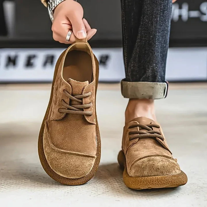Tan wide toe lace up shoe held up beside a worn shoe with rolled jeans