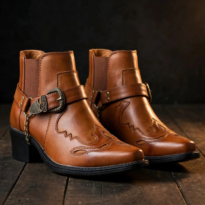 Pair of cognac harness ankle boots with buckle and chain on a wooden floor