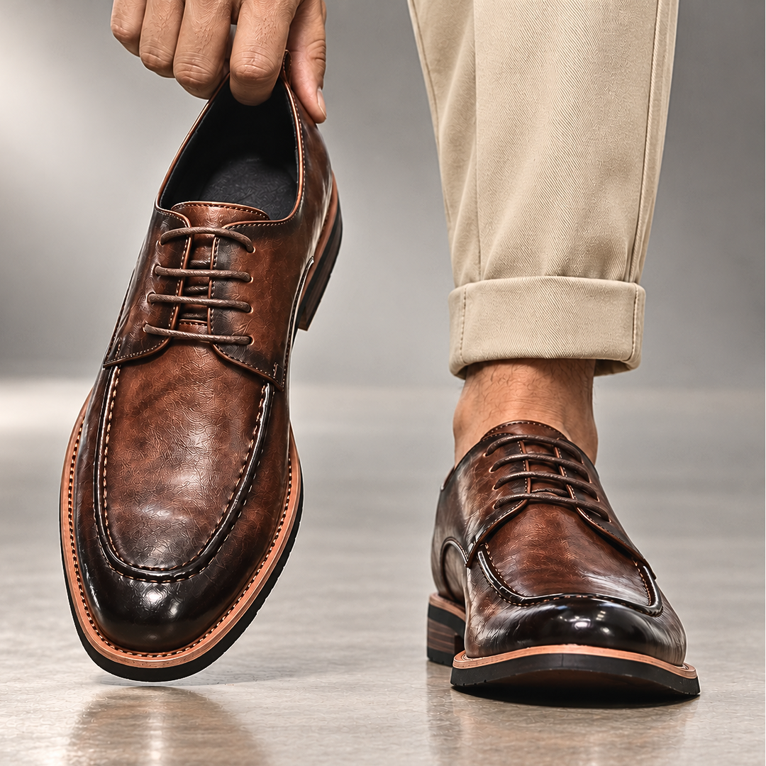 Hand lifting a brown lace up derby shoe beside beige cuffed trousers