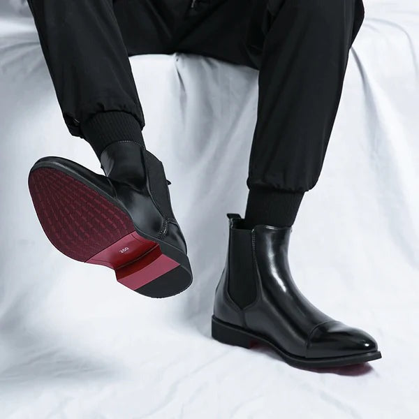 Black chelsea boot with lifted foot showing the red heel and sole on white fabric