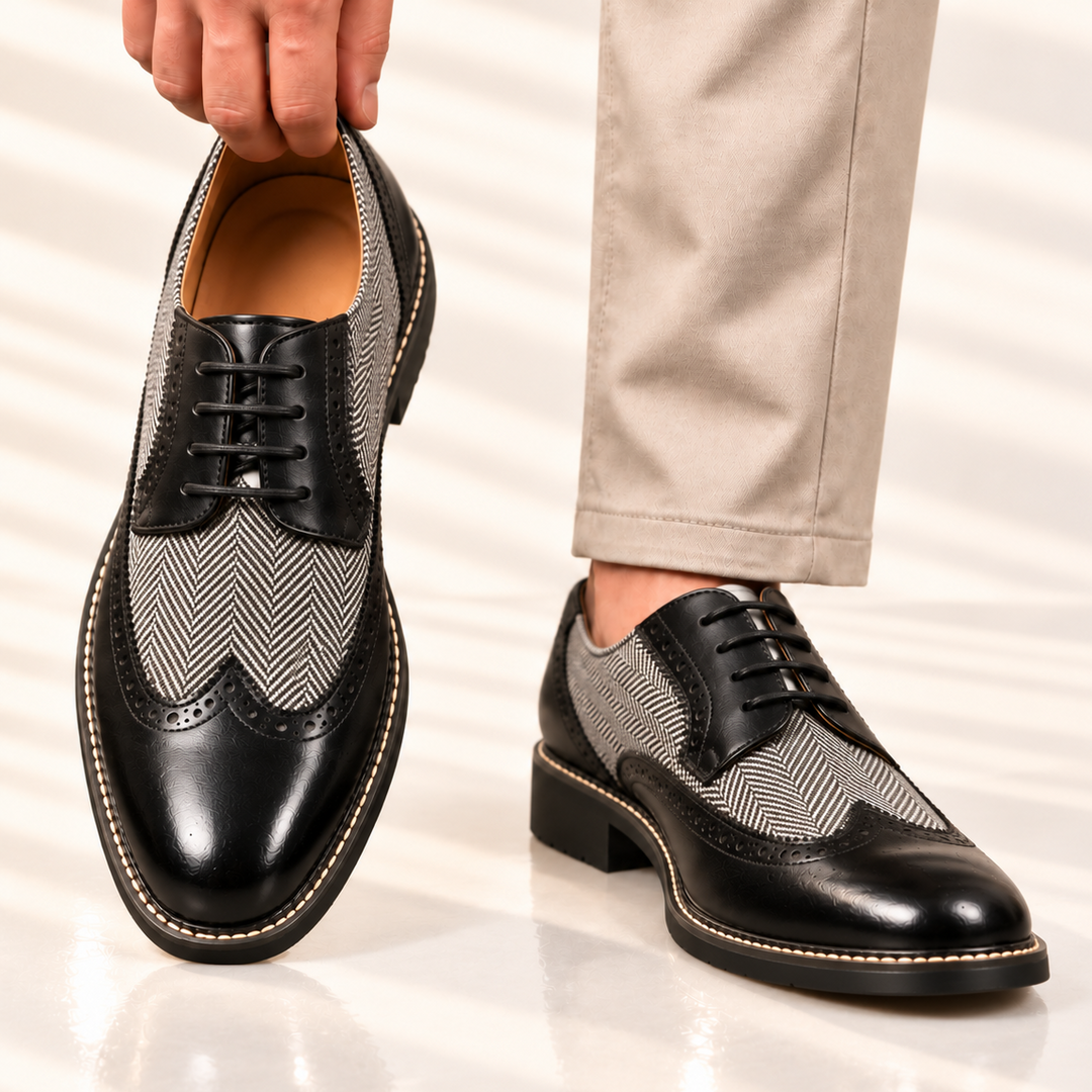Black wingtip brogue held in hand, with beige trousers and second shoe on foot