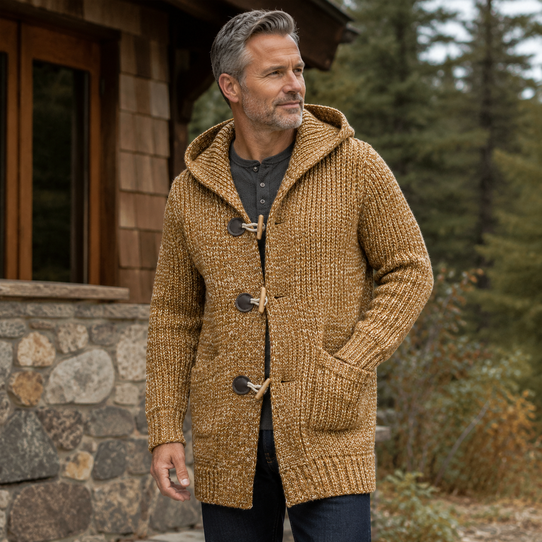 Man wearing the camel hooded cardigan outside a stone and wood cabin