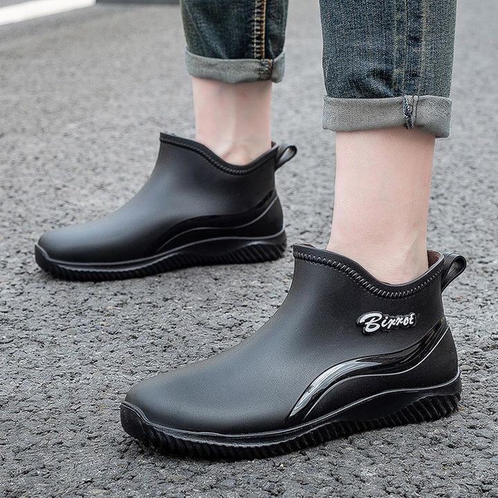 Black slip on rain boots worn outdoors with cuffed jeans, one boot in front