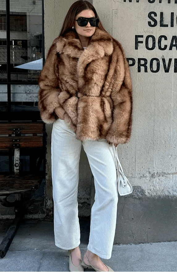 Model in the caramel short faux fur coat with white jeans standing outdoors