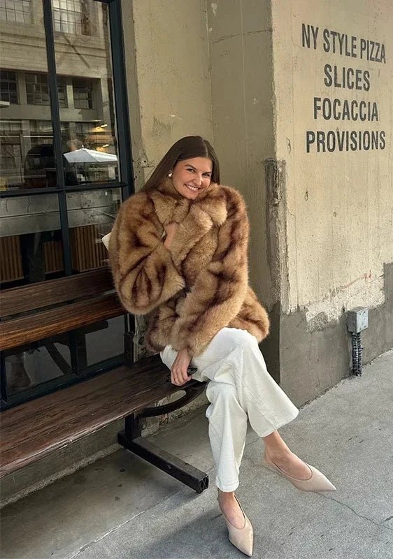 Model seated on a bench in the caramel short faux fur coat and white jeans