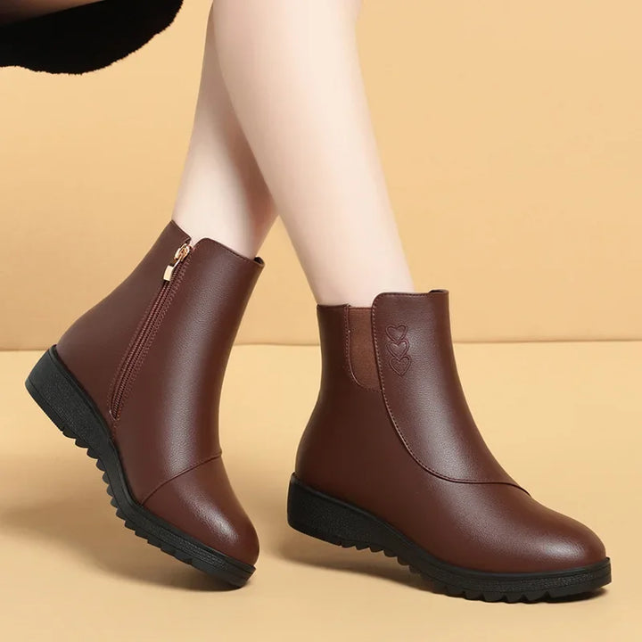 Brown round toe flat ankle boots with gold side zip, worn on a tan backdrop