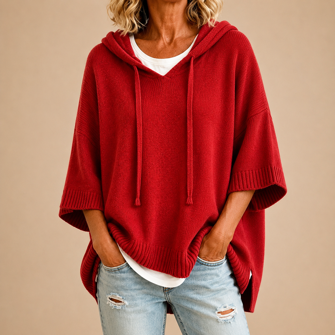 Red hooded knit poncho sweater with wide sleeves, hands in jean pockets