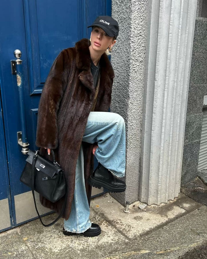 Model in the dark brown faux fur coat with a cap and blue jeans leaning against a doorway
