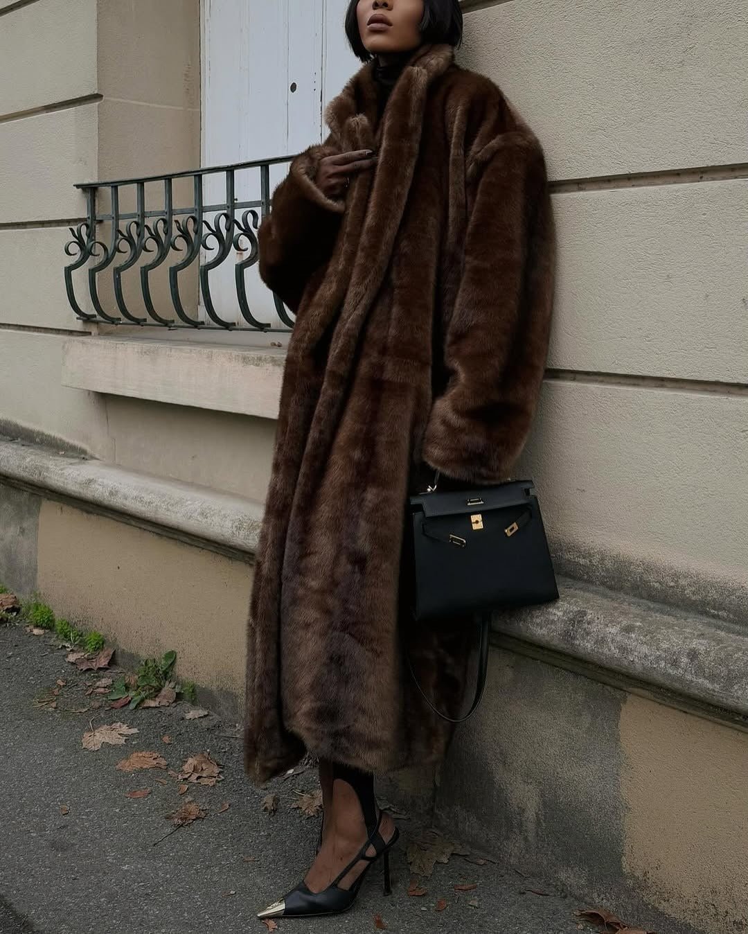 Model leaning on a wall in the dark brown maxi faux fur coat, long length to the ankle