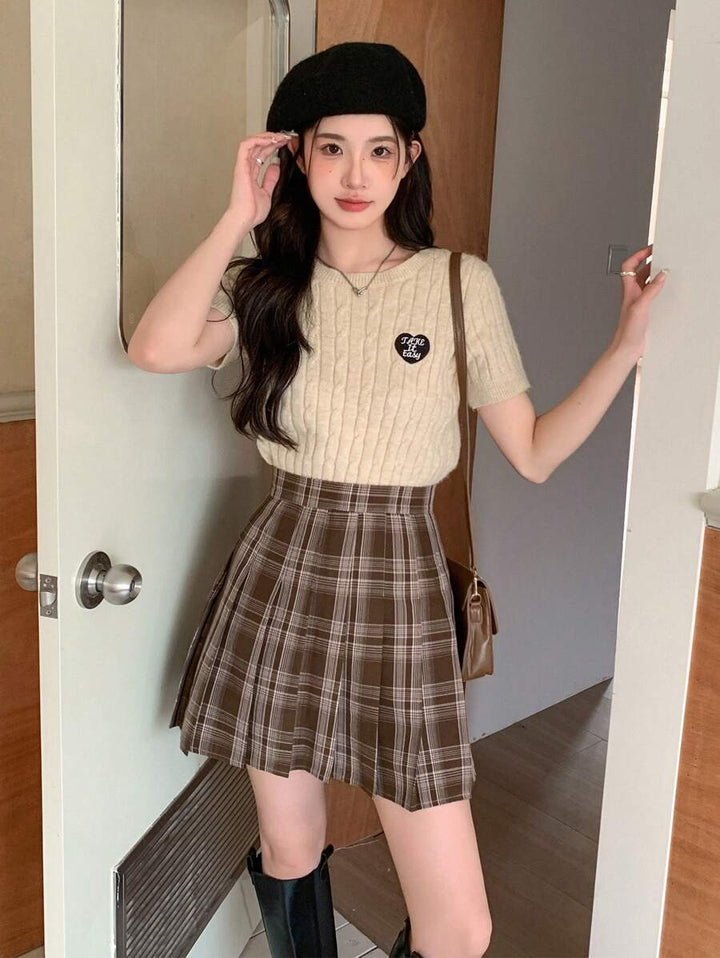 Model in a brown plaid skirt and cream knit top posing by a door