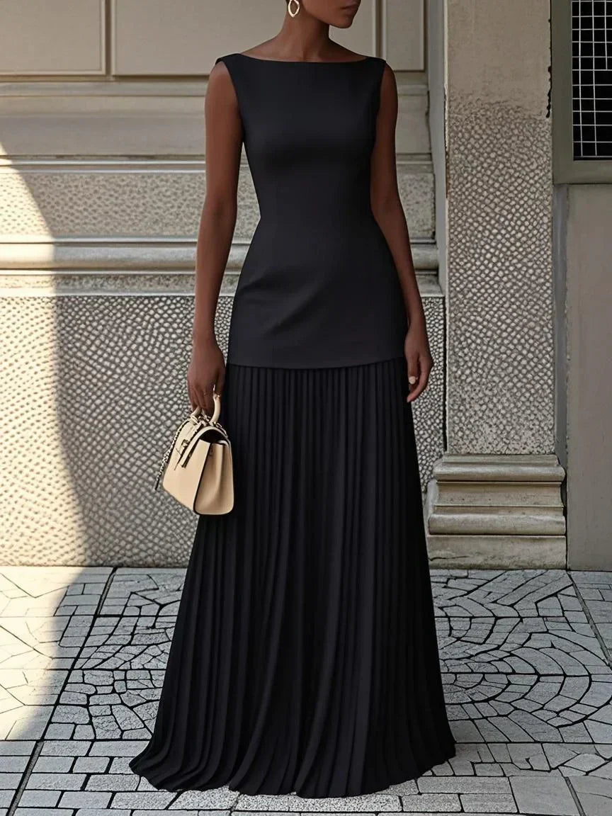 Black pleated maxi dress with boat neck and drop waist on a stone street