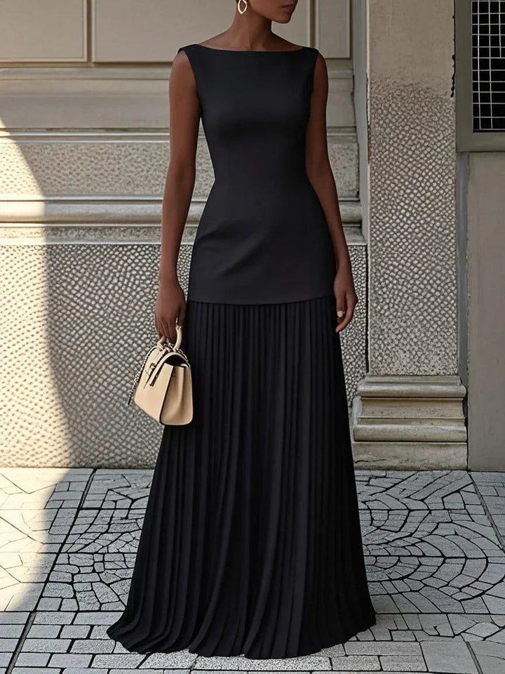 Black pleated maxi dress with boat neck and drop waist on a stone street