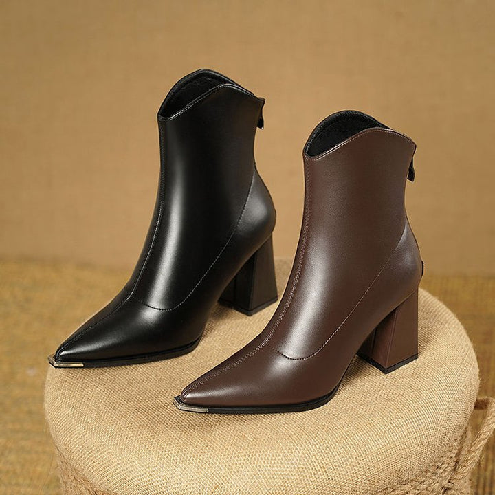 Black and brown pointed toe block heel ankle boots side by side on a woven stool