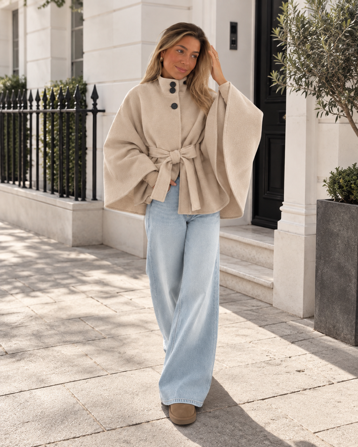 Beige belted poncho coat worn with light wide leg jeans on a sunny street