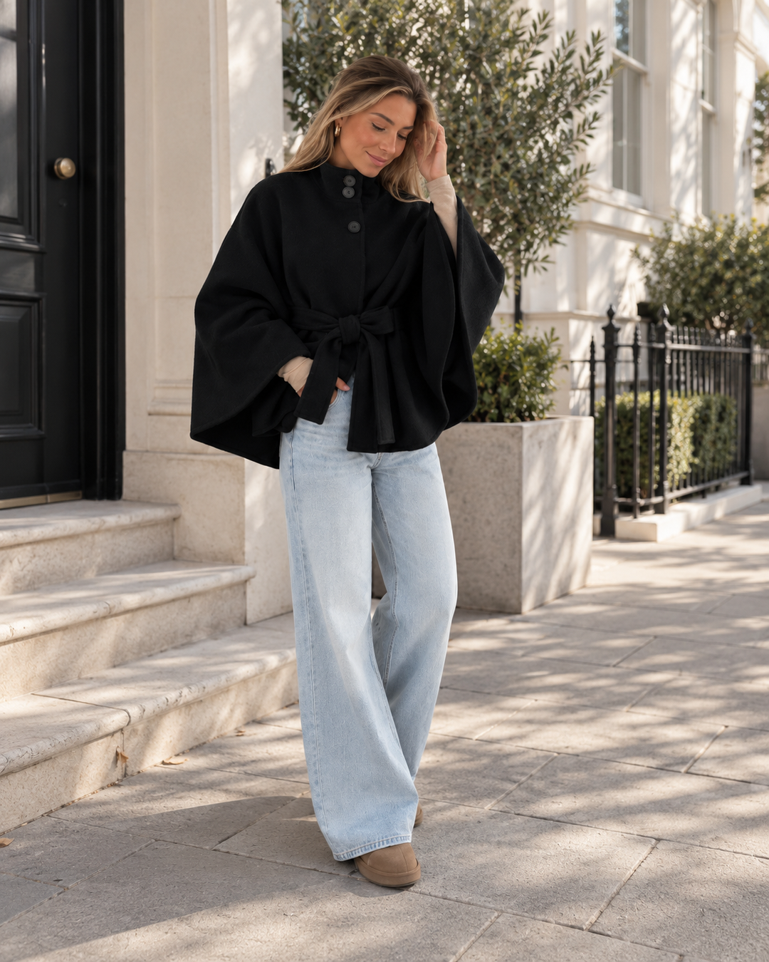 Black belted poncho coat styled with light blue wide leg jeans outside a townhouse