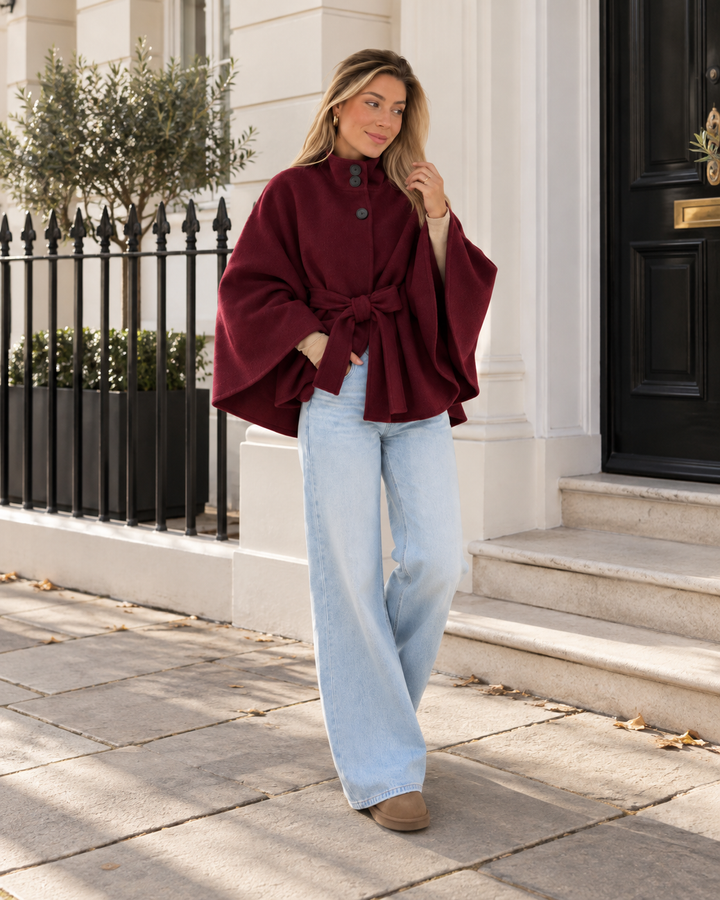 Burgundy belted poncho coat worn with wide leg jeans in front of a black door