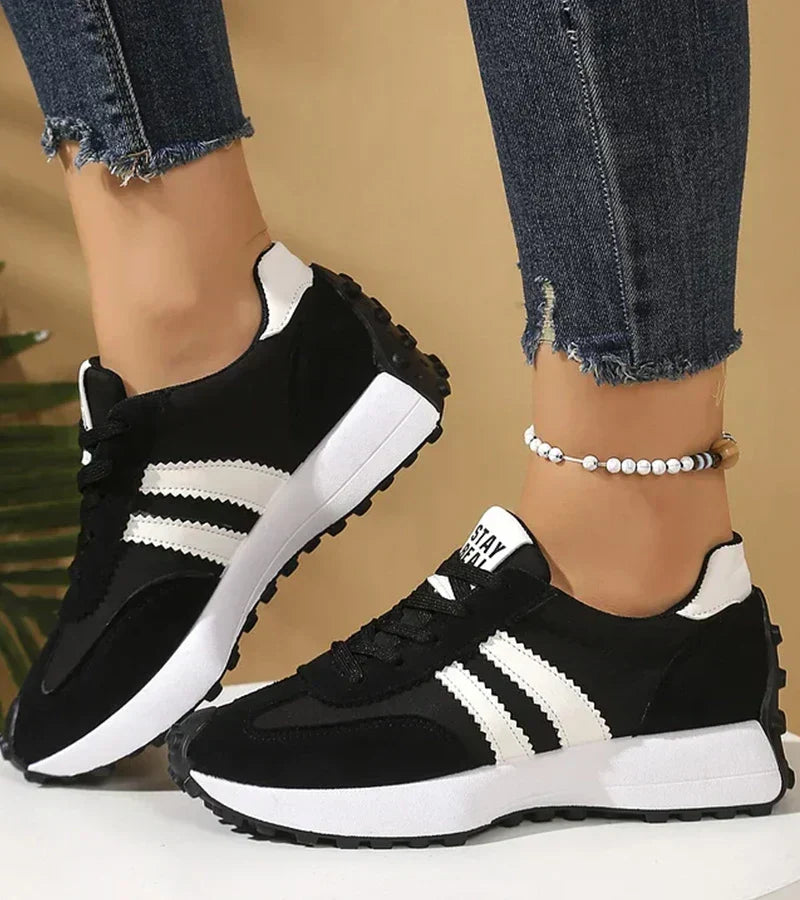 Black low top lace up sneakers with white stripes worn on a wooden floor