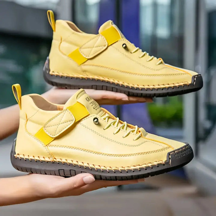 Pair of yellow stitched sneakers with strap, held out in hands outdoors
